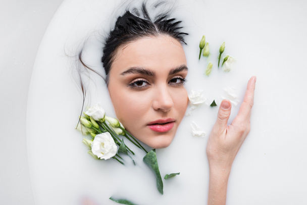 sensual mujer mirando a la cámara mientras toma baño de leche con flores eustoma - Foto, Imagen