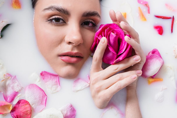 young woman looking at camera and holding rose while bathing in milk with floral petals - Foto, Imagen
