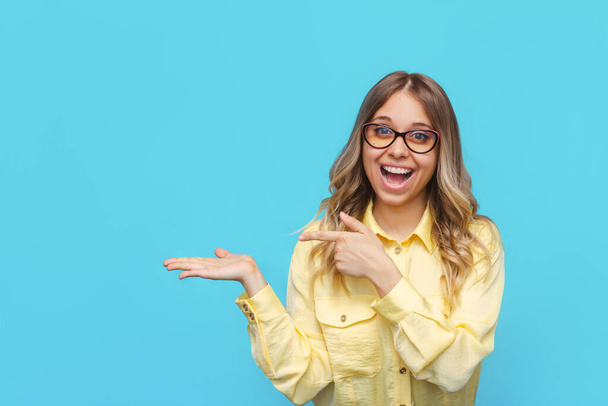 A young caucasian beautiful happy excited smiling blonde woman in a yellow shirt and glasses points with her index finger copyspace, hold hand demonstrating ads offer on a light color blue background - Foto, afbeelding