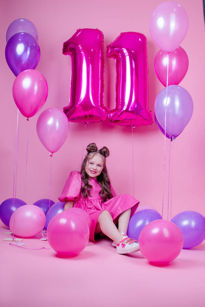 γλυκό καστανό κορίτσι με ροζ μακιγιάζ και σε ροζ δερμάτινο φόρεμα. κοντά πολύχρωμα μπαλόνια σε σχήμα έντεκα. σε ροζ φόντο.έννοια της χαράς, κόμμα, εορτασμός γενεθλίων - Φωτογραφία, εικόνα