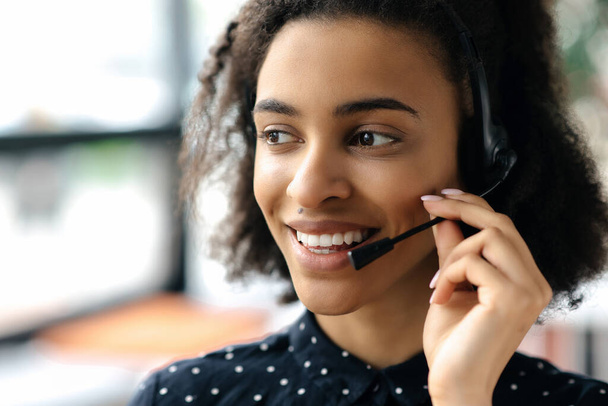 Close-up headshot of pleasant charming young african american curly haired woman in headset and stylish shirt, female call center worker or support operator, looking to the side, smiling friendly - Photo, Image