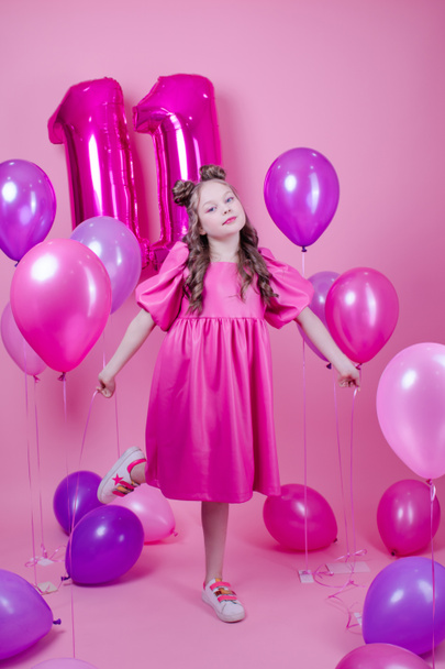 ピンクのメイクとピンクの革のドレスを着た甘いブルネットの女の子。11番の形をしたカラフルな風船の近くです。ピンクを背景に喜びパーティー誕生日のお祝い - 写真・画像