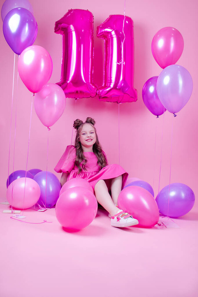 süßes brünettes Mädchen mit rosa Make-up und in rosa Lederkleid. Neben bunten Luftballons in Form der Zahl elf. auf rosa Hintergrund. Konzept der Freude, Party, Geburtstagsfeier - Foto, Bild