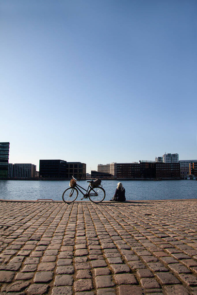 Blonde woman sitting on boardwalk and looking a the water in harbor with a bicycle standing next to her. Seen from behind and sun reflecting in water with cobblestones in the foreground. Copy space. - Foto, Imagem