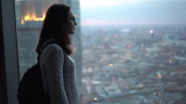 Young Asian woman with glasses looks out the window with a skyscraper in the evening. Girl in an apartment with panoramic windows. - Photo, Image