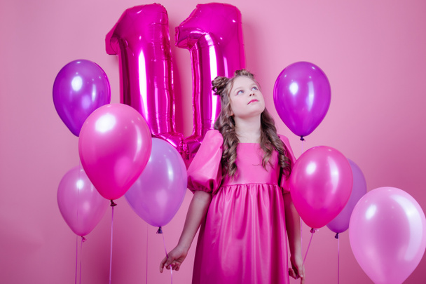 douce fille brune avec maquillage rose et en robe en cuir rose. près de ballons colorés en forme de numéro onze. sur fond rose. concept de joie, fête, célébration d'anniversaire - Photo, image