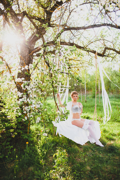 Young bride with blond hair in white negligee and stockings posing on a rope swing - Foto, Bild