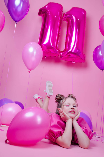 sweet brunette girl with pink make up and in pink leather dress. near colorful balloons in shape of number eleven. on pink background.concept of joy, party, birthday celebration - Fotó, kép