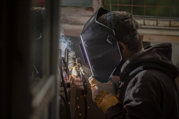 a welder in a welding mask, gloves welds metal with a welding machine and an electrode in his hand, sparks fly, a beautiful glow, bokeh, orange light on a black background, a flash of light and smoke - Фото, изображение