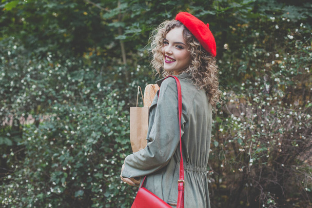 Mooie jonge vrouw in rode baret met ambachtelijke papieren zak met verse franse baguette tegen groene bladeren - Foto, afbeelding