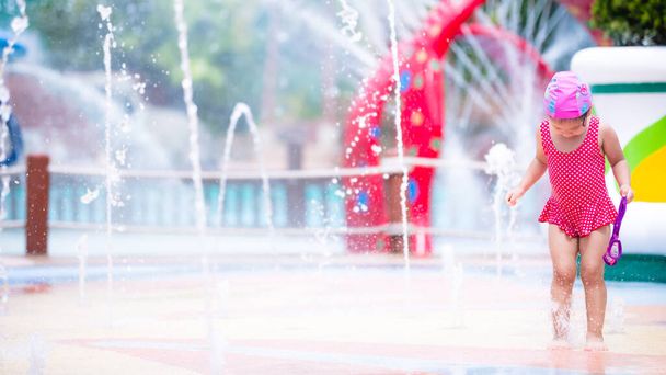Şirin Asyalı kız neşeyle yerden yükselen çeşmeye basıyor. Kırmızı mayo giyen çocuk yazın suda oynuyor. Mutlu bir çocukluk. Şehirdeki su parkındaki çeşme. 3-4 yaşındaki çocuk - Fotoğraf, Görsel
