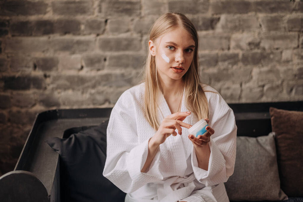Young blonde beautiful woman in the white bathrobe spread cream to her skin - Фото, изображение