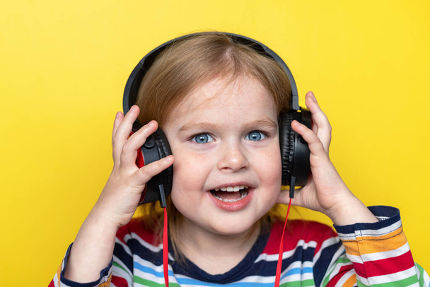 Happy smiling little child listening to music with large headphones, isolated on yellow background. - 写真・画像