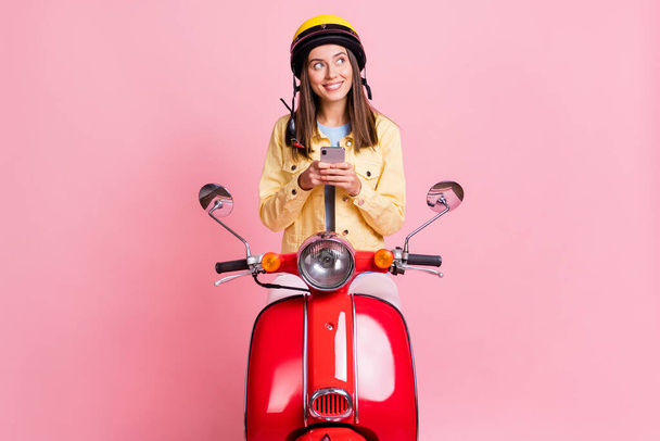 Photo of young curious happy girl look copyspace hold phone wear yellow helmet ride red motorbike isolated on pink color background - Фото, зображення