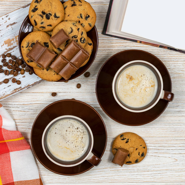オープンブックを背景にチョコレートチップクッキーとチョコレートとコーヒーの2つのカップ。ティールームでのチョコレートチップクッキーとホットコーヒー時間. - 写真・画像