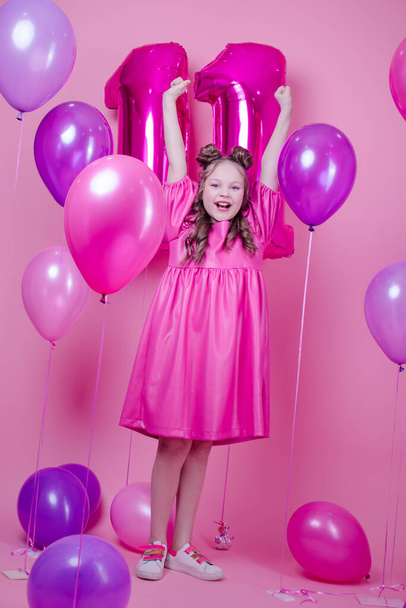 сладкая брюнетка девушка с розовым макияжем и в розовом кожаном платье. около красочных шариков в форме числа одиннадцать. на розовом фоне. Концепция радости, вечеринки, празднования дня рождения - Фото, изображение