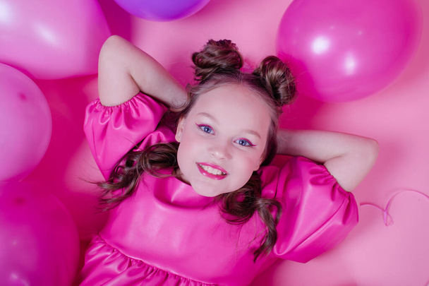 ピンクのメイクとピンクの背景にカラフルな風船の近くのピンクの革のドレスで魅力的なブルネットの女の子のモデル。喜び、パーティー、誕生日の概念 - 写真・画像
