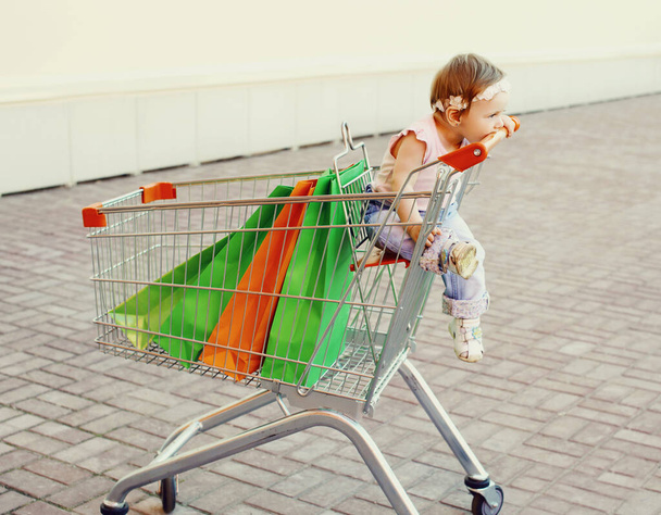 Happy baby with shopping bags sitting in a trolley cart in a city - Foto, Imagen