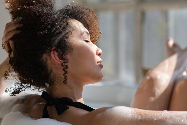 back view curly black girl laying, relaxing in the bath indoors. young woman relaxing in warm bathtub with foam and bubbles. Tired female enjoying rest pamper herself. - Fotoğraf, Görsel