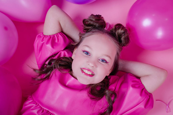 vonzó barna lány modell rózsaszín smink és rózsaszín bőr ruha közelében színes lufik rózsaszín hátteren.koncepció öröm, party, születésnap - Fotó, kép