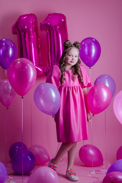 сладкая брюнетка девушка с розовым макияжем и в розовом кожаном платье. около красочных шариков в форме числа одиннадцать. на розовом фоне. Концепция радости, вечеринки, празднования дня рождения - Фото, изображение