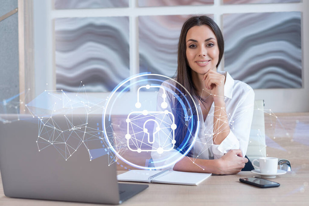 Attractive businesswoman in white shirt at workplace working with laptop to defend customer cyber security. Concept of clients information protection. Padlock hologram over office background. - Fotó, kép