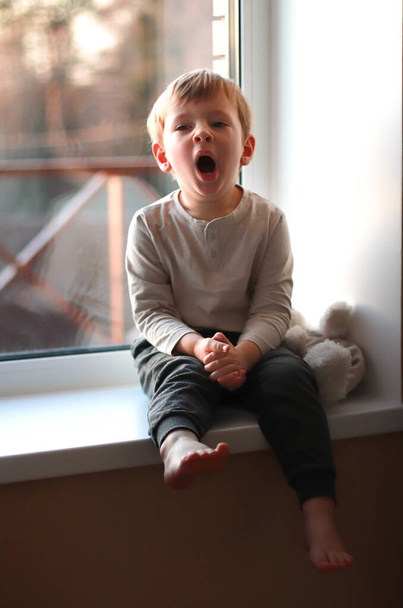Um menino se senta em um peitoril da janela na luz da noite com ursinho de pelúcia e bocejos. Dia Internacional da Criança. - Foto, Imagem