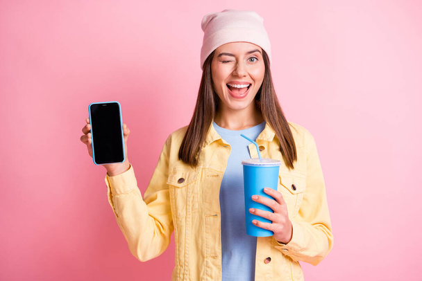 Portrait of attractive person hold drink demonstrate phone eye wink headwear cap isolated on pink color background - 写真・画像