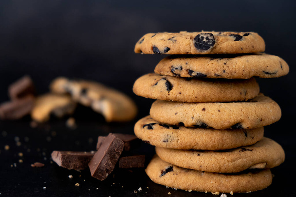 Chocolate chip cookies stack on black background. Recipe of homemade biscuits with pieces of chocolate. Dessert menu presentation for restaurant or cafe. Delicious cookies with chocolate crumbs. - 写真・画像
