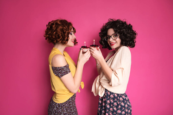 Két aranyos lány barátok nő néz ki, mint kocka könyvelők álló rózsaszín háttér. Göndör barna parókát és ízléstelen retro alkalmi ruhákat viselnek. Bogyós süti gyertyával a kezükben.. - Fotó, kép