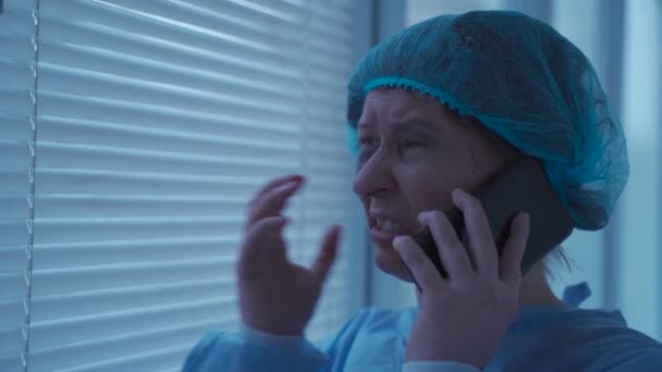 Θλιβερή γυναίκα ασθενής με ιατρικά ρούχα, μιλάει στο τηλέφωνο και κλαίει μόνη της στο νοσοκομείο, γυναίκα γιατρός αναμονής στο παράθυρο του νοσοκομείου, αισθάνεται στρεσαρισμένη, κλαίει και κινητό στο smartphone λόγω ατυχήματος - Πλάνα, βίντεο
