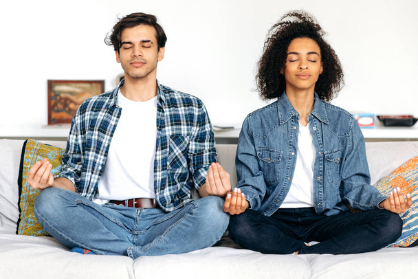 Uvolněný multiraciální zamilovaný manželský pár, sedící v lotosové pozici na gauči doma v neformálním oblečení, uvolňující napětí se zavřenýma očima, meditující, muž a dívka spolu tráví čas - Fotografie, Obrázek
