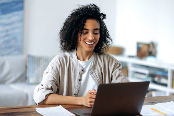 Joyful cute confident curly haired stylish african american girl, in casual wear, freelancer or student working remotely, chatting with colleague or classmate via video call, friendly smiling - Photo, image