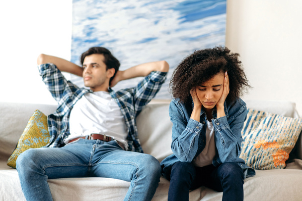 A split in family relationships. Disgruntled hispanic guy and upset african american girl sitting on the couch and not talking to each other after quarrel, experiencing stress and frustration - Fotó, kép