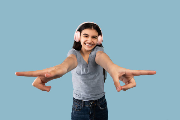 Indian teen girl in casual wear wearing wireless headphones, pointing fingers at different sides on blue background - Fotó, kép