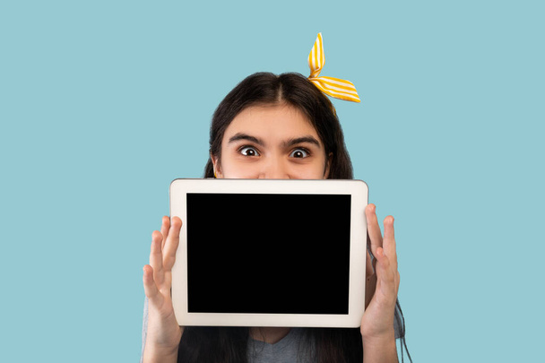 Adolescente india escondida detrás de la tableta PC con pantalla en blanco sobre fondo azul, espacio para el diseño del sitio web o aplicación - Foto, Imagen