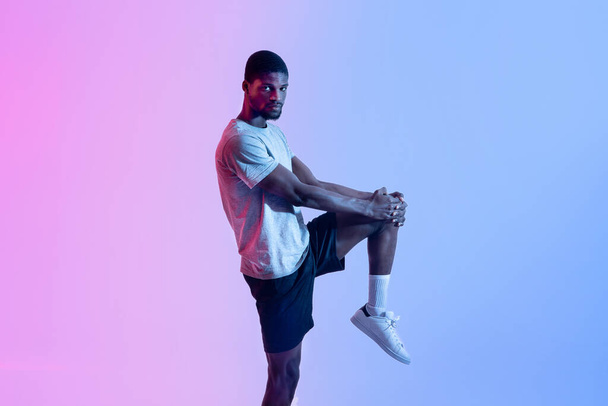 Portret wysportowanego czarnego mężczyzny rozciągającego nogę w neonowym oświetleniu. Koncepcja zdrowego stylu życia - Zdjęcie, obraz