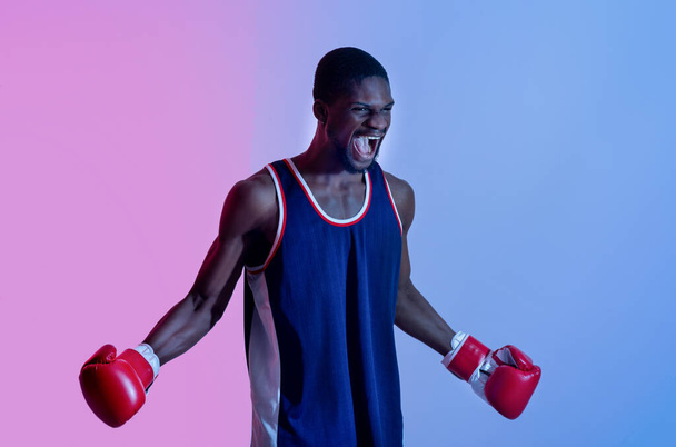 Агрессивный чёрный спортсмен в боксёрских перчатках кричит в гневе в неоновом свете. Концепция боевого спорта - Фото, изображение