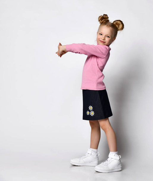 Smiling blonde girl with a stylish hairstyle, in a pink blouse with a throat and a stylish skirt with daisy flowers and sneakers posing looking at the camera - Fotó, kép