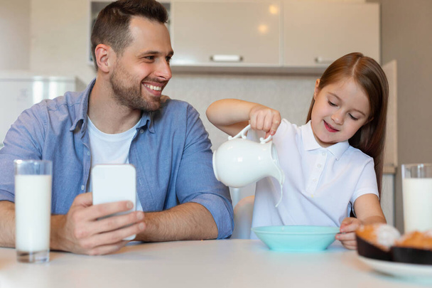 Χαρούμενος πατέρας και κόρη προσθέτοντας γάλα στα δημητριακά στην κουζίνα - Φωτογραφία, εικόνα