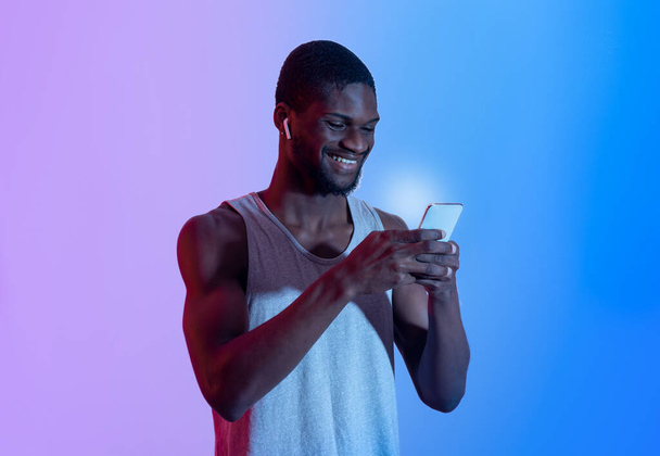 Attractive African American guy with cellphone wearing earphones, picking music playlist in neon light - Photo, Image