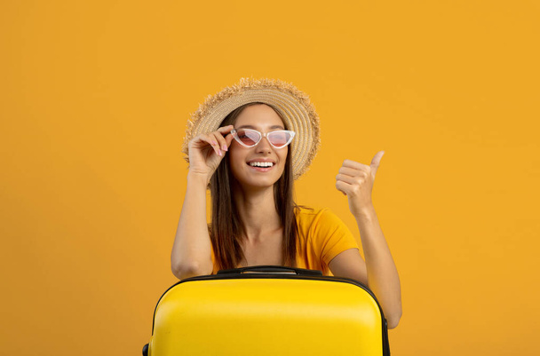 Весела жінка в сонцезахисних окулярах спирається на валізу, показуючи простір копіювання
 - Фото, зображення