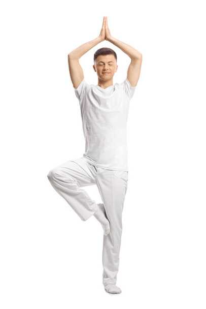 Full length portrait of a young man in white clothes practicing yoga and standing on one leg isolated on white background - Fotó, kép
