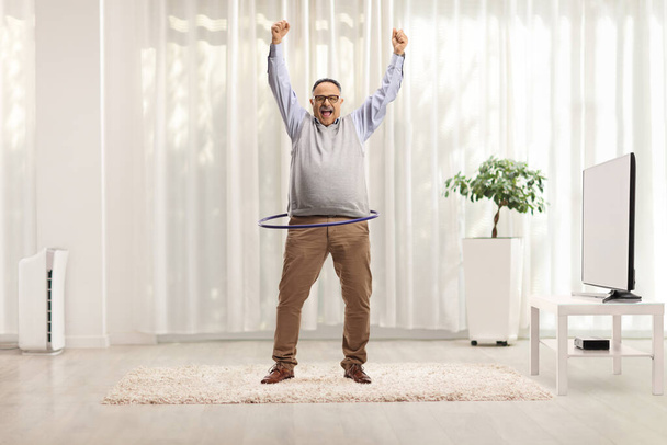 Full length portrait of a happy mature man spinning a hula hoop and lifting arms up in a living room - Фото, зображення