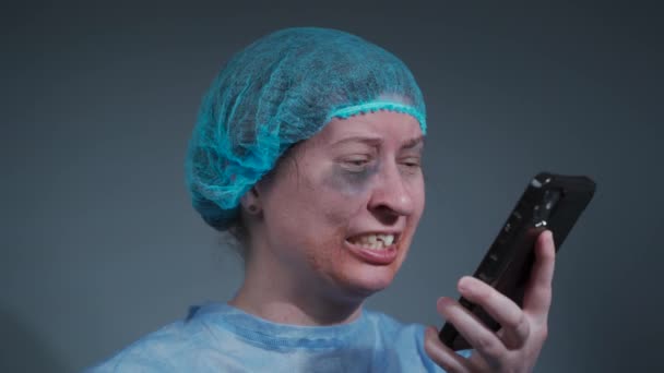 Wanhopige vrouw in blauwe beschermende operatiejurk met gezicht, gebit verwondingen na ongeval wacht op een operatie in het ziekenhuis, huilt mobiele telefoon, brengt slecht nieuws aan familie in de eerste hulp. Verzekeringsoproep - Video