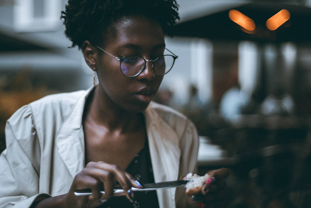A low-key portrait of a young beautiful black woman in eyeglasses and white trench, and with nail art, sitting in an evening restaurant and spreading butter or cheese on a piece of bread - Photo, Image