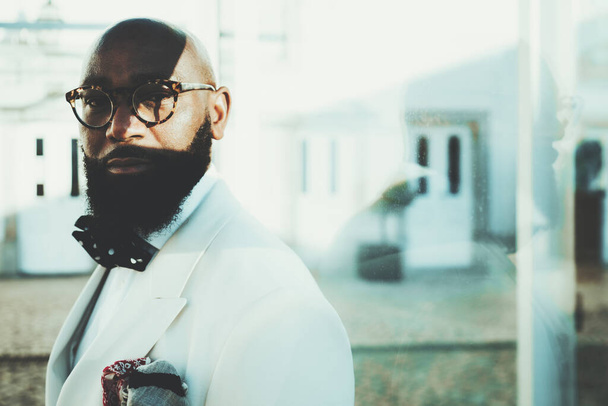 Portrait of an elegant stately bald bearded black guy in eyeglasses and a fashionable white custom-made costume with dotted bow-tie, standing on the street with a contrast shadow on a part of the face - Fotografie, Obrázek