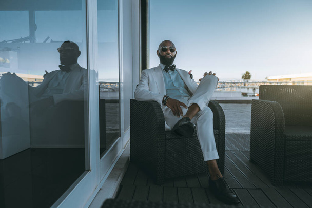 A smart stately black guy in sunglasses and a white costume with a dotted bow tie is sitting with legs crossed on a wicker armchair outdoors on a restaurant terrace near the glass wall, warm evening - Foto, afbeelding
