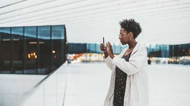 Side view of a young beautiful elegant black female in glasses, white trench coat, and with nail art, taking pics in a gallery of a modern business office building using a camera of her cellphone - Фото, зображення