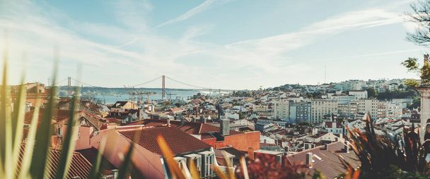 Panoramic shot of a sunny Lisbon cityscape from the high above; the panorama of Lisbon urban landscape with plenty of antique and modern houses and a suspension bridge over Tagus river in a background - Zdjęcie, obraz